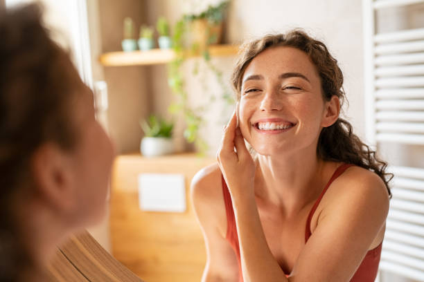 Cheerful young woman using cotton pad while looking in mirror. Happy smiling beautiful girl cleaning skin with cotton pad. Beauty natural woman looking in mirror while cleansing skin face and using cosmetic products for properly deep clean.